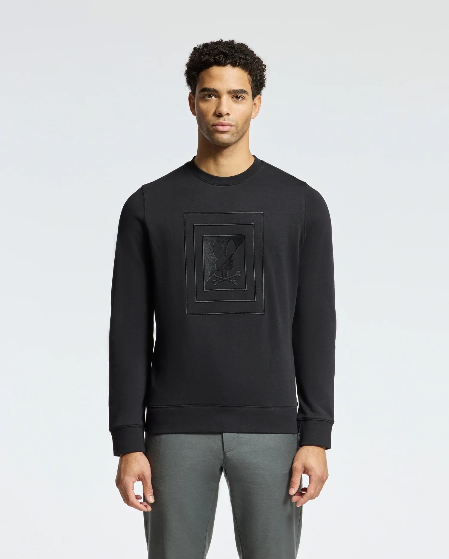 A man stands against a plain light gray background, wearing the Psycho Bunny MENS OAK CITY CREW NECK - B6S274E200 in black, featuring subtle raised embroidery on the front. His short curly hair complements his style as he effortlessly pairs this premium quality piece with gray pants.