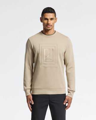 A person wearing the MENS OAK CITY CREW NECK - B6S274E200 sweater by Psycho Bunny, featuring raised embroidery on the chest in beige, stands against a plain white background, paired with black pants.