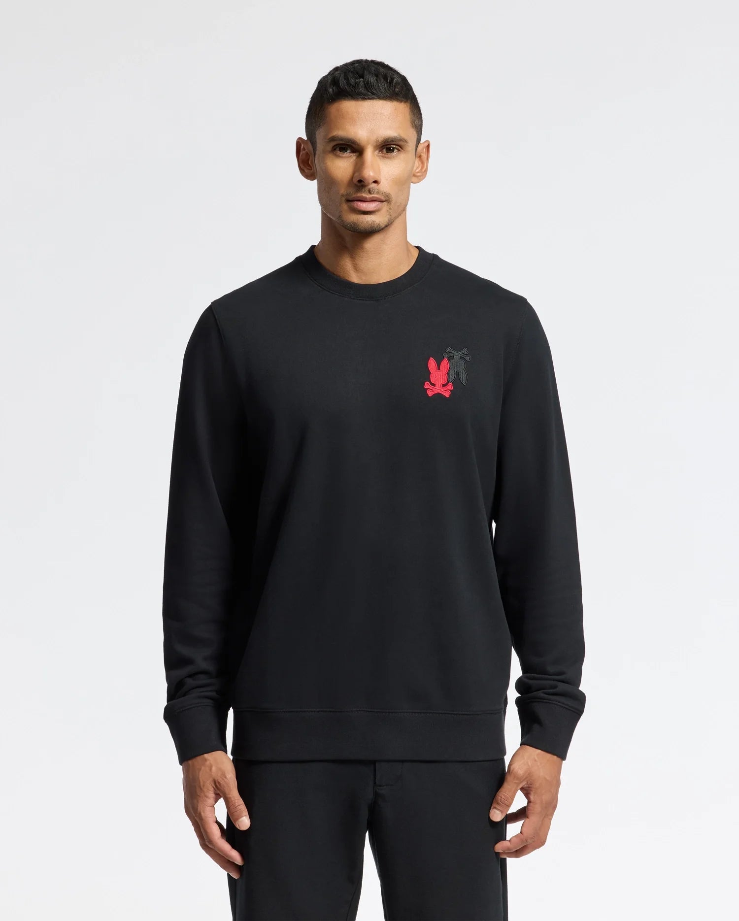 A man is wearing the Psycho Bunny MENS PIERCE PEACHED TERRY CREW - B6S278E200, a black sweatshirt with a small red graphic on the chest. He stands against a plain white background.