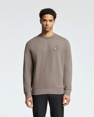 A man stands against a light gray background wearing the Psycho Bunny MENS PIERCE PEACHED TERRY CREW, B6S278E200. It's a stylish cotton crew with a small cartoon graphic on the chest. He has short, curly hair and pairs it with sleek black pants.