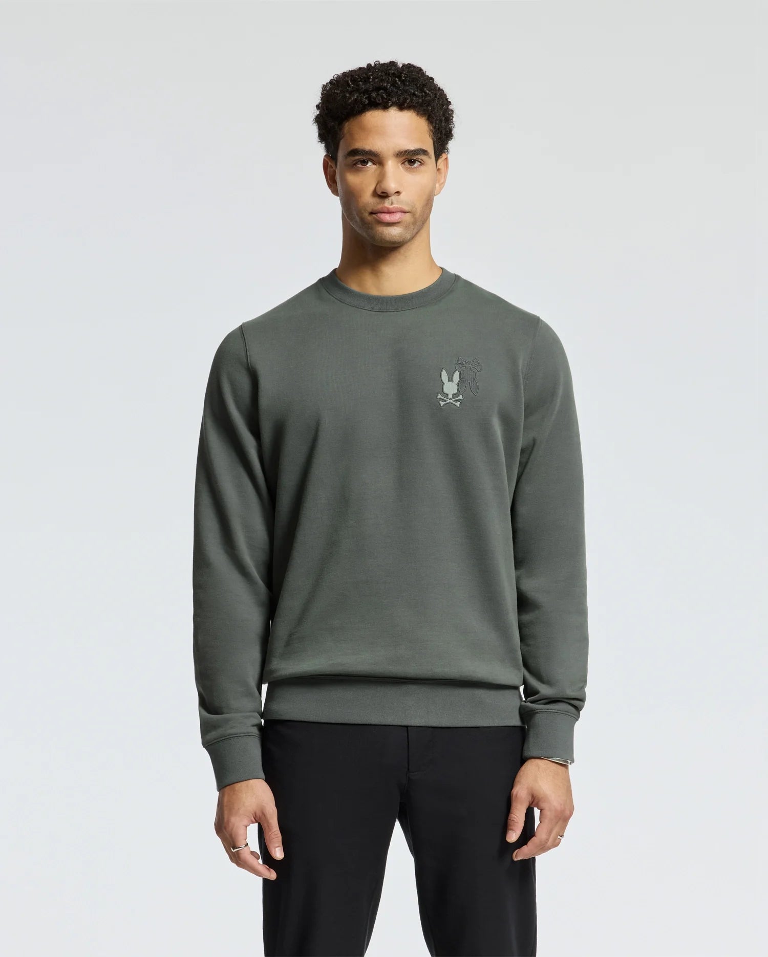 A person with short curly hair is wearing a dark green MENS WORDMARK PEACHED TERRY CREW - B6S278E200 by Psycho Bunny, featuring an embroidered bunny logo on the left chest. They are standing against a plain gray background.