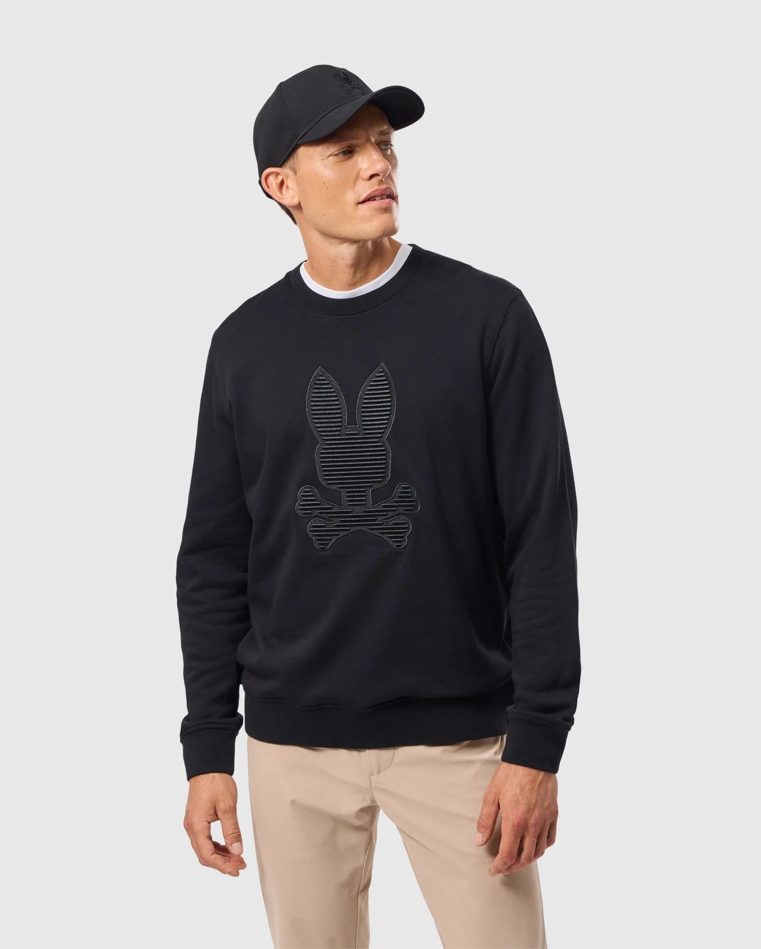 A man stands against a plain background, wearing a Psycho Bunny MENS KINGSTON SWEATSHIRT - B6S480D200 with a 3D-embroidery bunny and crossbones design. He pairs this black cotton jersey top with a white undershirt and beige pants, thoughtfully looking to the side.