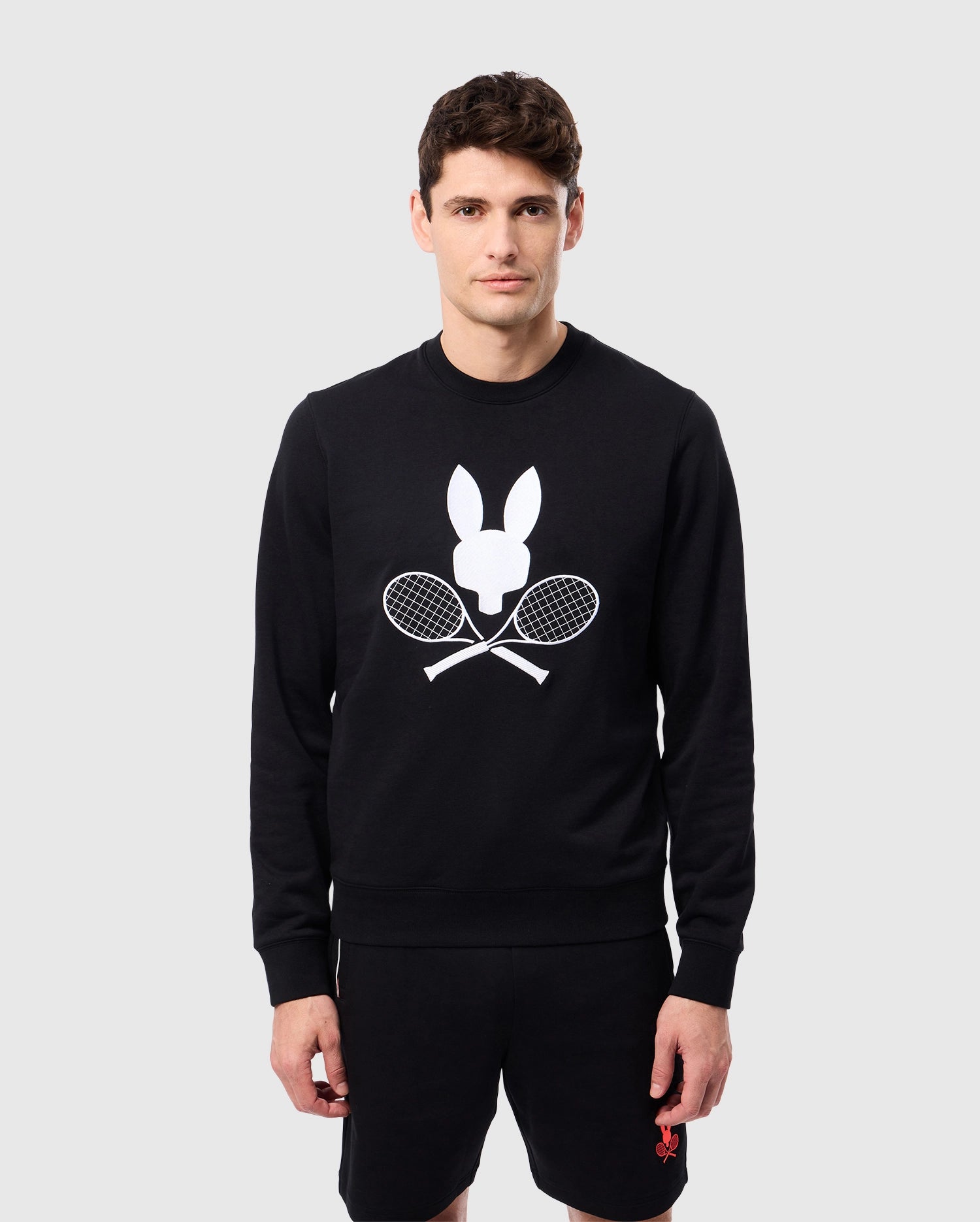 A man wears the MENS COURTSIDE SWEATSHIRT by Psycho Bunny, showcasing a white bunny emblem with crossed tennis rackets. He pairs it with black shorts against a plain light gray background, exuding a tennis-inspired style.