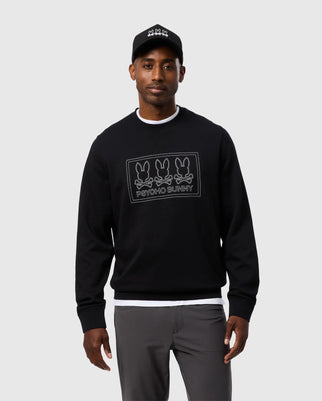 A man stands against a light gray background, wearing the Psycho Bunny MENS TACOMA MODAL SWEATSHIRT - B6S927D200 in black, featuring premium modal fabric and adorned with a graphic of three bunnies and text, paired with gray pants.