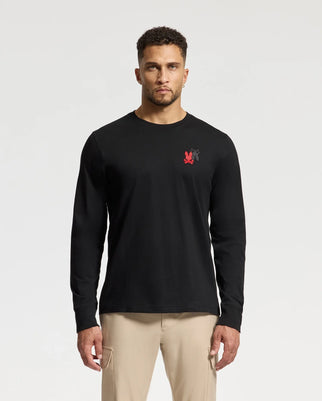 A man stands against a plain background wearing the Psycho Bunny MENS PIERCE LONG SLEEVE BACK GRAPHIC TEE - B6T178E200, which is made from black Pima cotton and features a subtle red and black graphic on the chest. He pairs it effortlessly with beige pants for a sleek look.