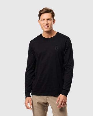 A person wearing a Psycho Bunny MENS ALBERTON LONG SLEEVE TEE (B6T867D200) in black Pima cotton with a subtle emblem on the left chest area and beige pants stands against a plain light background. They are smiling, and their hair is neatly styled.