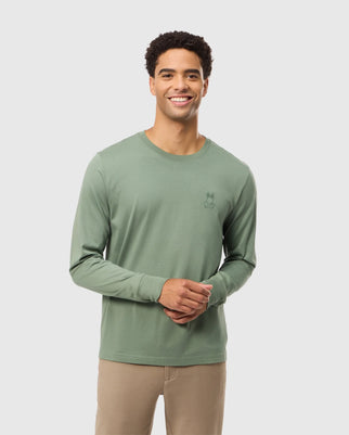 A person with short, curly hair is smiling while wearing a soft Pima cotton sage green MENS ALBERTON LONG SLEEVE TEE (B6T867D200) by Psycho Bunny and beige pants. The shirt features a minimalist graphic of a cactus and the word "friendly" on the left chest. The background is neutral grey.