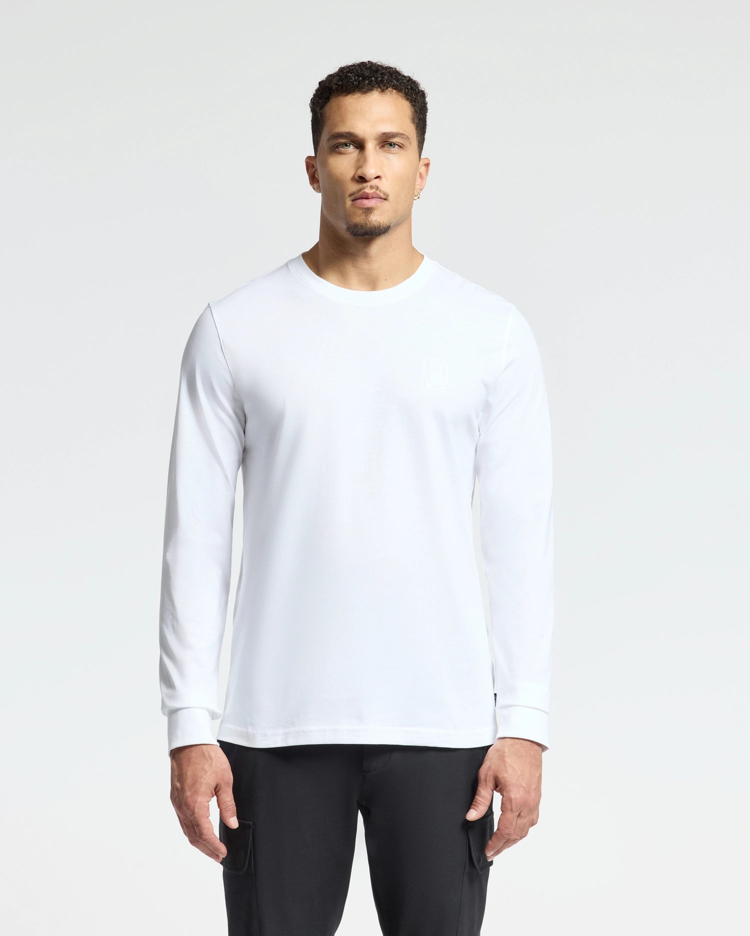 A person stands against a plain white background, wearing a Men's Alberton Long Sleeve Tee by Psycho Bunny and black pants. Their arms are relaxed by their sides, facing the camera with a neutral expression.