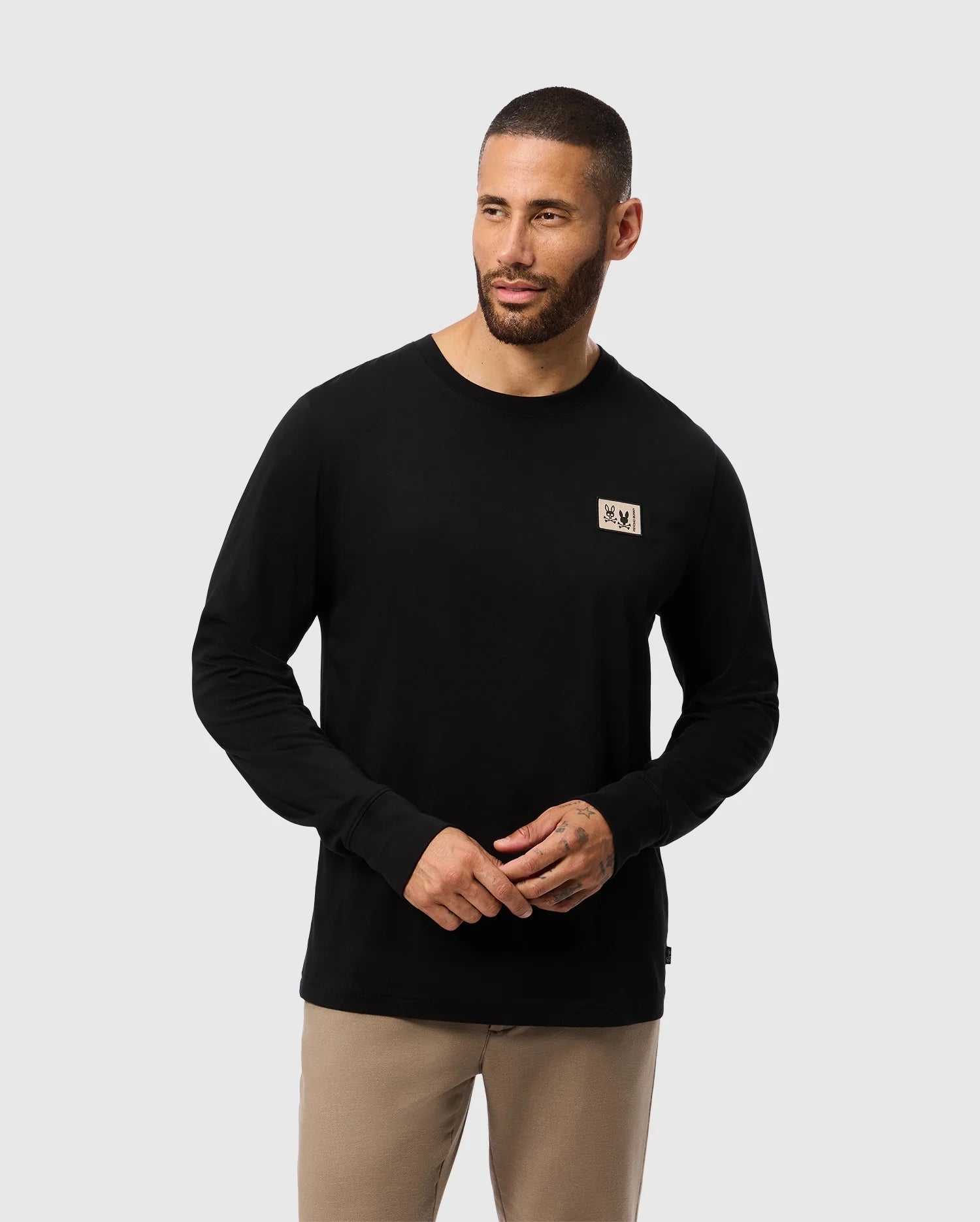 A bearded man wears the Psycho Bunny MENS MONROE LONG SLEEVE TEE - B6T975D200 in black, crafted from pima cotton and adorned with a small chest patch. Complementing the beige pants, this regular fit outfit exudes a versatile, cozy style. He looks to the side against a plain light background.