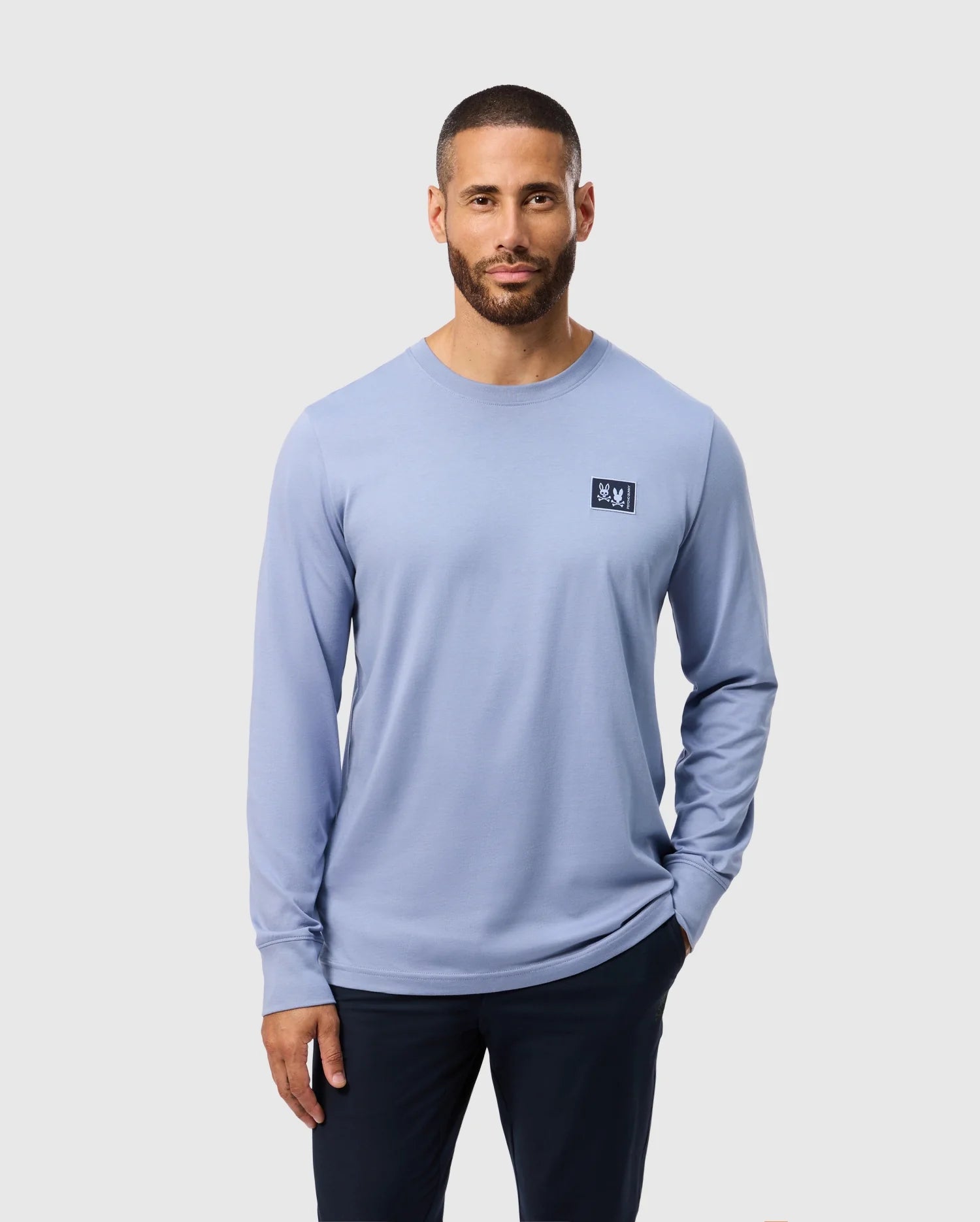 A man with short hair and a beard is wearing the Psycho Bunny MENS MONROE LONG SLEEVE TEE in light blue, crafted from lightweight and durable Pima cotton, featuring a small logo on the chest. He pairs it with dark pants and stands against a plain gray background, hands in his pockets.