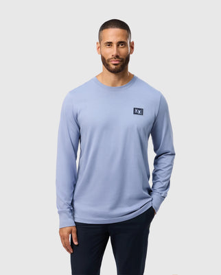 A man with short hair and a beard is wearing the Psycho Bunny MENS MONROE LONG SLEEVE TEE in light blue, crafted from lightweight and durable Pima cotton, featuring a small logo on the chest. He pairs it with dark pants and stands against a plain gray background, hands in his pockets.