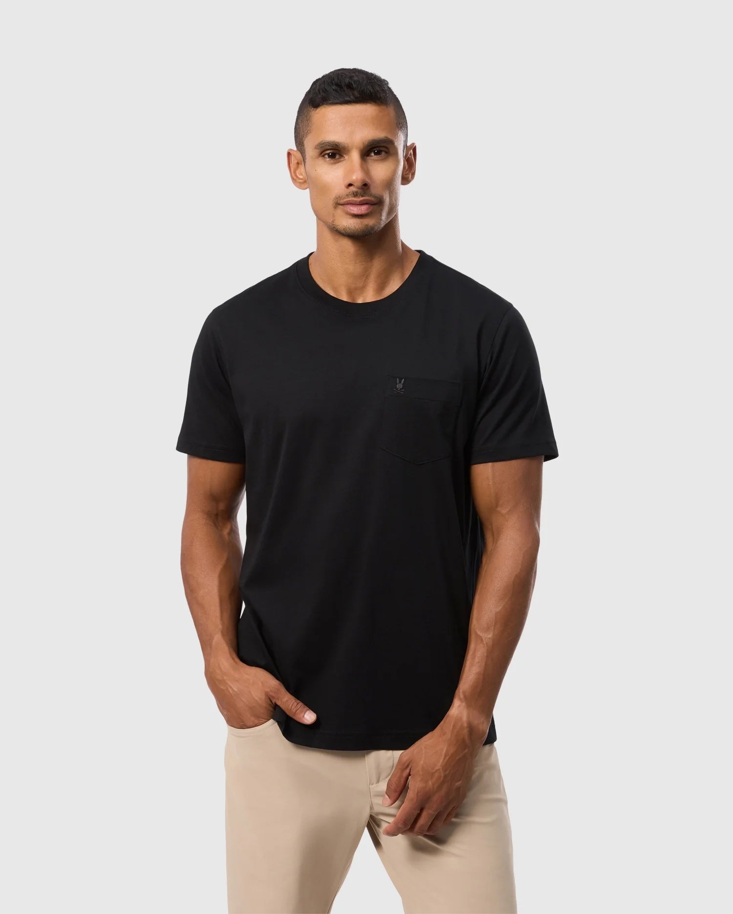 A person with short dark hair wears Psycho Bunny's MENS BONNIE REGULAR FIT POCKET TEE in black, crafted from 100% Pima cotton and made in Peru, paired with beige pants. A small pocket adorns the left side of the T-shirt. The backdrop is a plain gray color.