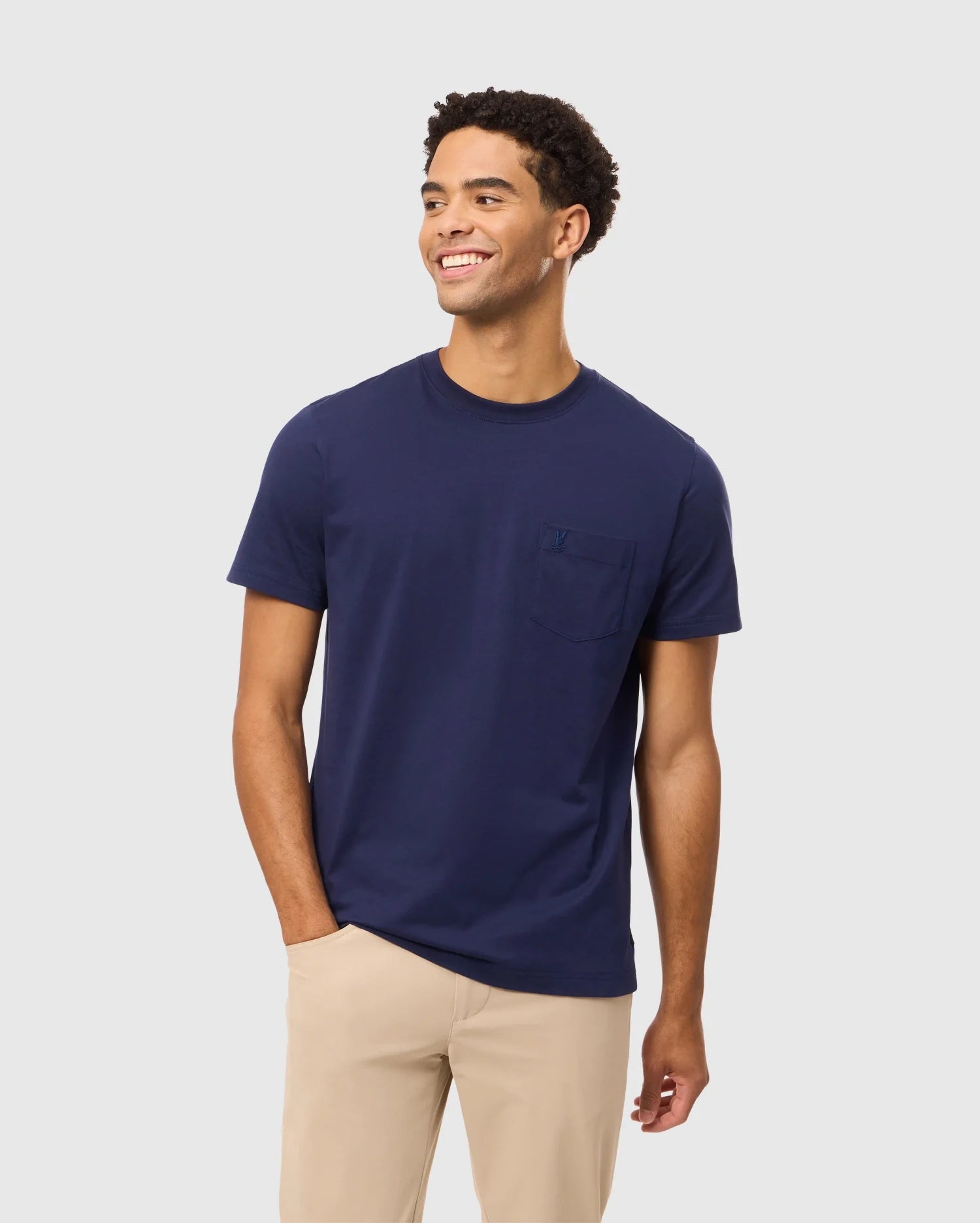 A person in a navy blue Psycho Bunny MENS BONNIE REGULAR FIT POCKET TEE - B6U011D200 with an Arrow chest pocket, made from Pima cotton, stands against a plain light gray background, smiling and looking to the side while wearing beige pants.