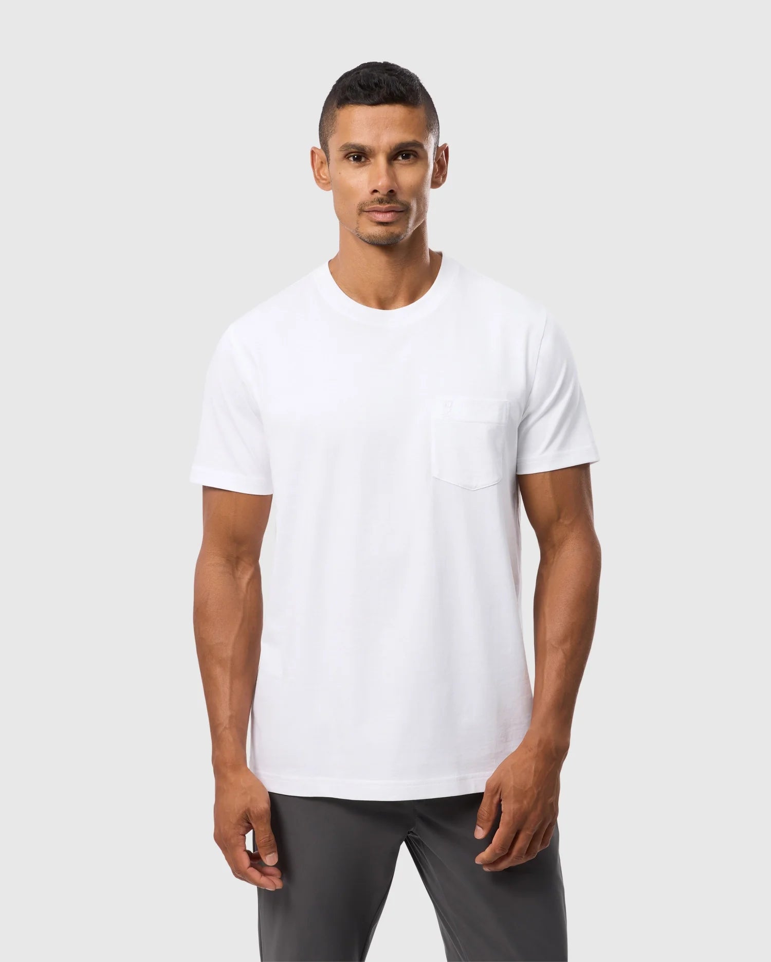 A person with short hair stands neutrally against a plain background, wearing a Psycho Bunny MENS BONNIE REGULAR FIT POCKET TEE in white, made from soft Pima cotton, paired with dark gray pants.