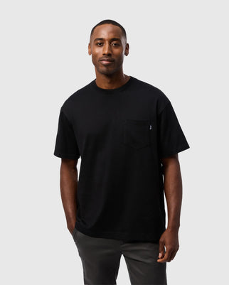 Wearing a relaxed-fit Psycho Bunny Men's Clyde Oversized Pocket Tee - B6U012D200 in black Pima cotton and dark pants, a person stands against a plain gray background, smiling gently at the camera.