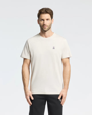 A man with short hair and a beard is wearing Psycho Bunny's MENS CLASSIC CREW NECK TEE - B6U014B200, made from premium Pima cotton jersey. The white tee has a small embroidered logo on the chest and is paired with black pants. He stands against a neutral background, facing the camera.