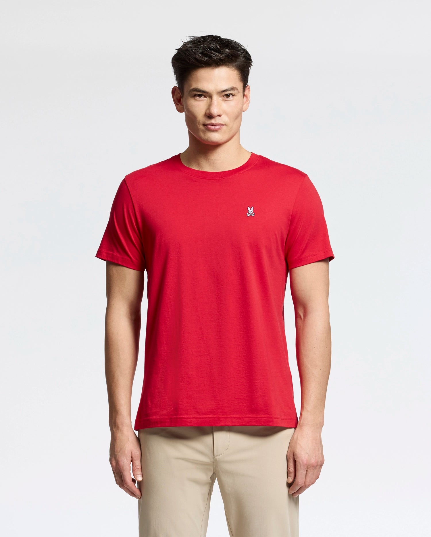 A man wearing a red Psycho Bunny MENS CLASSIC CREW NECK TEE (B6U014CRPC) with a small embroidered logo on the left chest stands against a white background, paired with beige pants. He has short, dark hair.