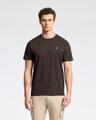 A man wears a Psycho Bunny MENS CLASSIC CREW NECK TEE crafted from premium Pima cotton in plain black with a small logo on the left chest, paired with light beige pants, standing against a neutral white background.