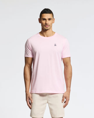 A man stands facing forward wearing a Psycho Bunny Men's Classic Crew Neck Tee in light pink pima cotton, featuring a small chest logo, paired with beige shorts against a plain white background.