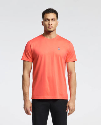 A man stands against a white background wearing Psycho Bunny's MENS CLASSIC CREW NECK TEE (B6U014F200) in bright coral, crafted from soft Pima cotton with an embroidered Bunny logo on the chest, paired with black pants.