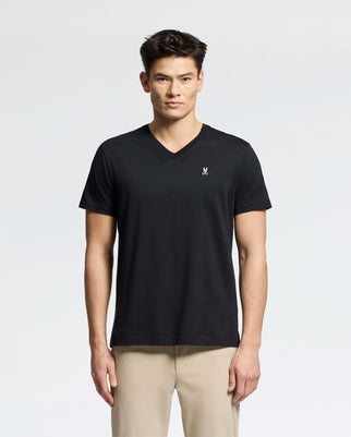 A person in a black Psycho Bunny MENS CLASSIC V NECK TEE - B6U100ARPC, made of soft Pima cotton with a small white logo on the chest, stands against a plain background. They have short dark hair and are dressed in beige pants.