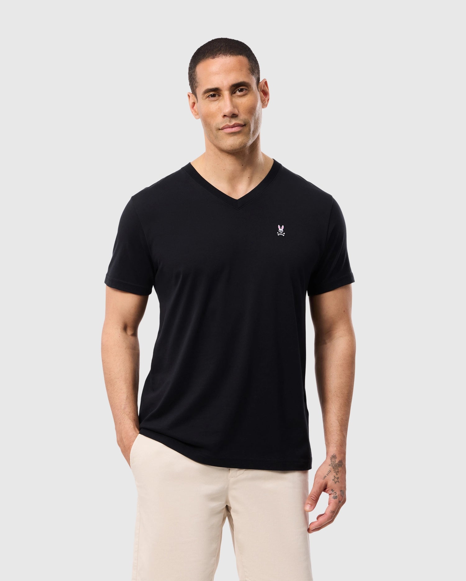 A man wearing a black Psycho Bunny MENS CLASSIC V NECK TEE - B6U100ARPC made from soft Pima cotton, along with beige pants, stands against a plain gray background. He has short hair and a thoughtful expression, with one hand in his pocket and a tattoo visible on his left hand.