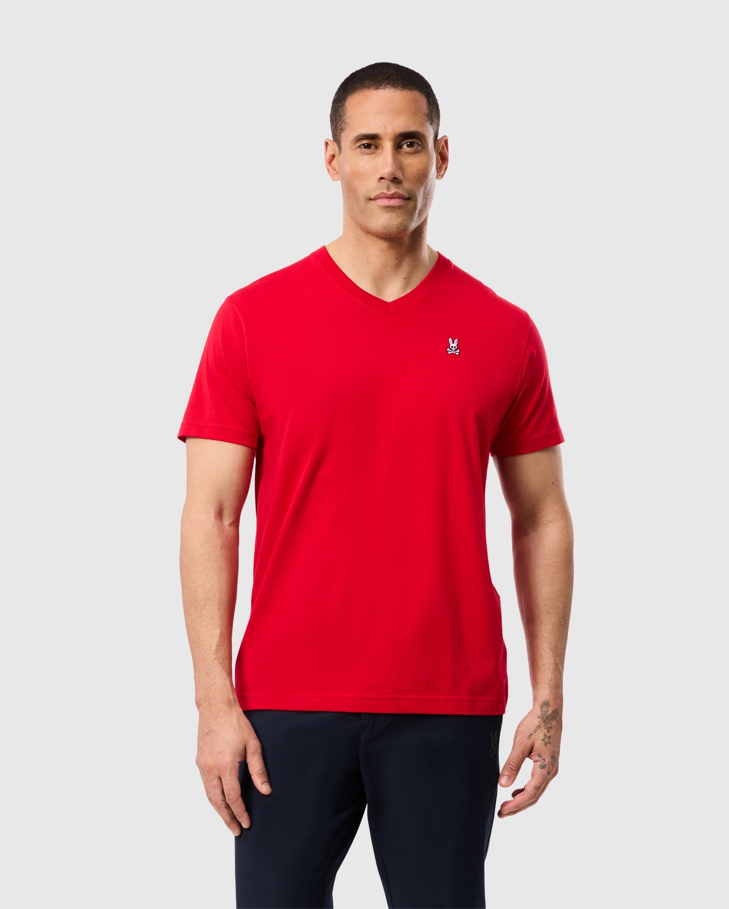 A man with short hair is wearing a bright red Psycho Bunny MENS CLASSIC V NECK TEE - B6U100CRPC, made from 100% Pima cotton, featuring a small white embroidered Psycho Bunny logo on the left chest. He is also wearing dark pants and stands against a plain light gray background.