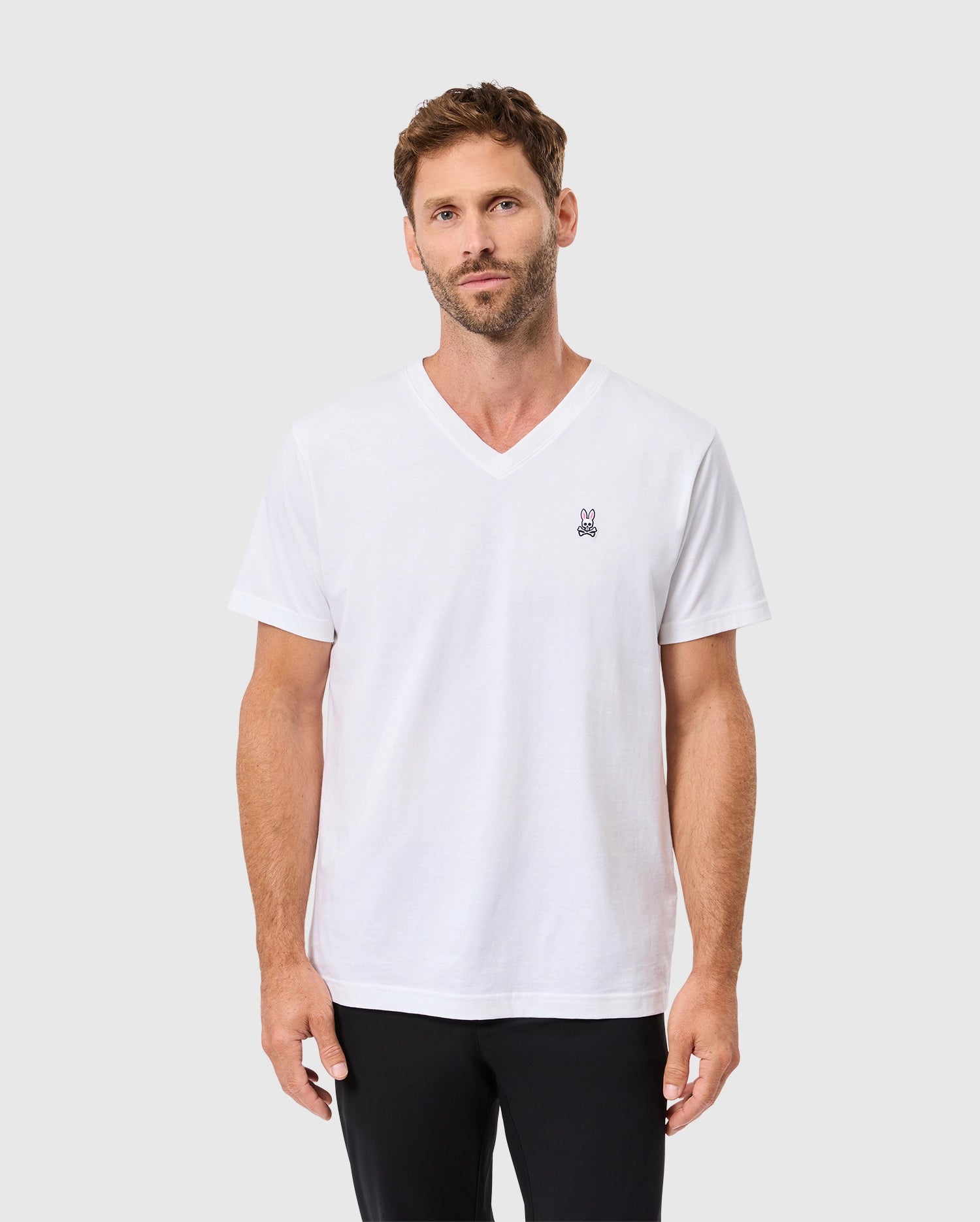 A man with short brown hair and a beard wears the MENS CLASSIC V NECK TEE - B6U100ARPC by Psycho Bunny, made from 100% Pima cotton. The plain white T-shirt is adorned with a small black embroidered logo on the left chest as he stands against a plain white background.