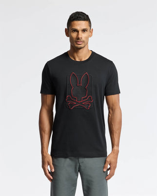 A man wearing a Psycho Bunny MENS PIERCE EMBROIDERED GRAPHIC TEE - B6U115E200, which showcases a red outlined bunny and crossbones design on Pima cotton, stands against a plain white background. He has short, dark hair and is wearing gray pants.