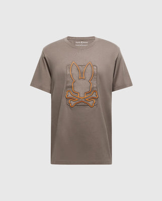 The Psycho Bunny MENS PIERCE EMBROIDERED GRAPHIC TEE - B6U115E200 features a stylish brown short-sleeve design made from premium Pima cotton, adorned with a striking orange-outlined bunny embroidery above two crossed bones, elegantly centered on the chest against a plain white background.