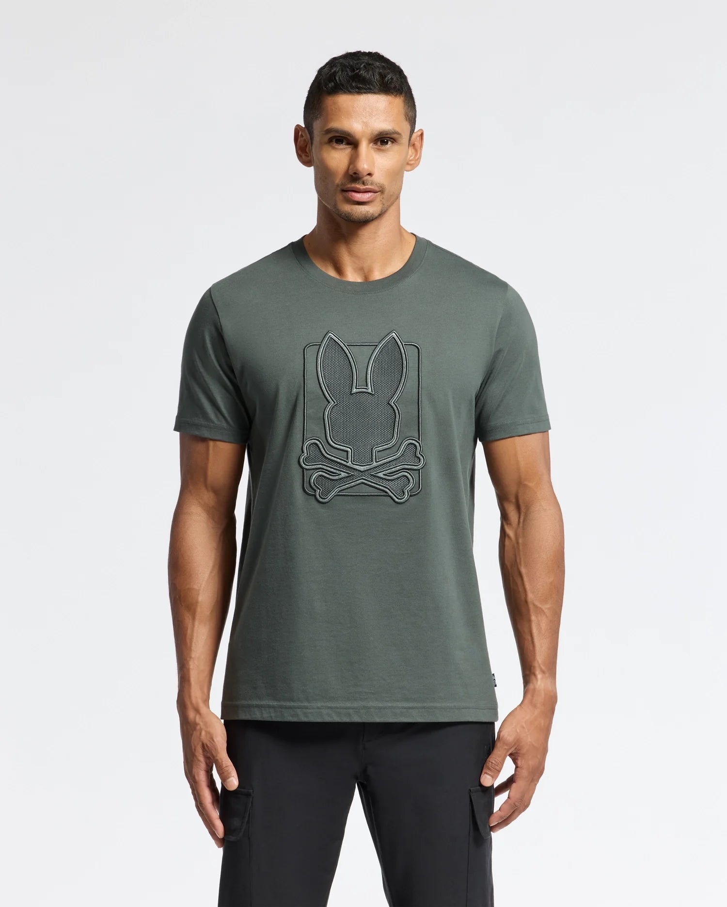 A man wearing a dark green Psycho Bunny MENS PIERCE EMBROIDERED GRAPHIC TEE - B6U115E200, made from soft Pima cotton, features a prominent emblem with bunny ears and crossed bones. He stands against a plain white background, paired with sleek black pants.