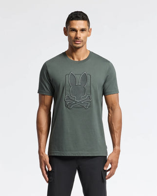 A man wearing a dark green Psycho Bunny MENS PIERCE EMBROIDERED GRAPHIC TEE - B6U115E200, made from soft Pima cotton, features a prominent emblem with bunny ears and crossed bones. He stands against a plain white background, paired with sleek black pants.