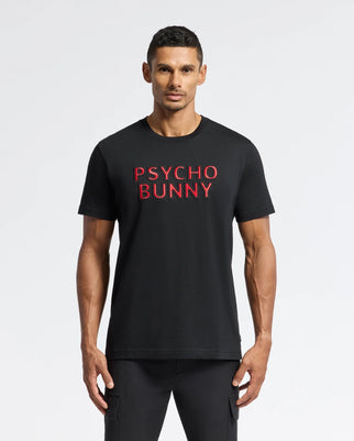 A man stands wearing the MEN'S PIERCE EMBROIDERED GRAPHIC TEE - B6U116E200 by Psycho Bunny, featuring "Psycho Bunny" embroidered in red on the front. He has short, dark hair and is facing forward against a plain white background, highlighting this stylish graphic tee.