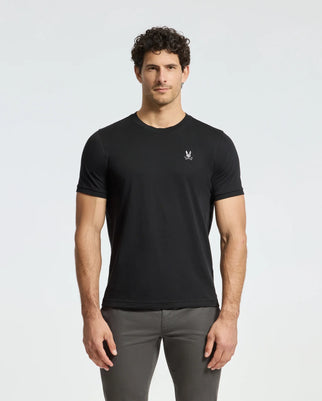 A man with curly hair wears a fitted black Psycho Bunny Men's Alexander Fashion Tee (B6U139E200) featuring a small logo on the left chest, paired with dark gray pants. He stands against a plain, light gray background, facing forward with a neutral expression.