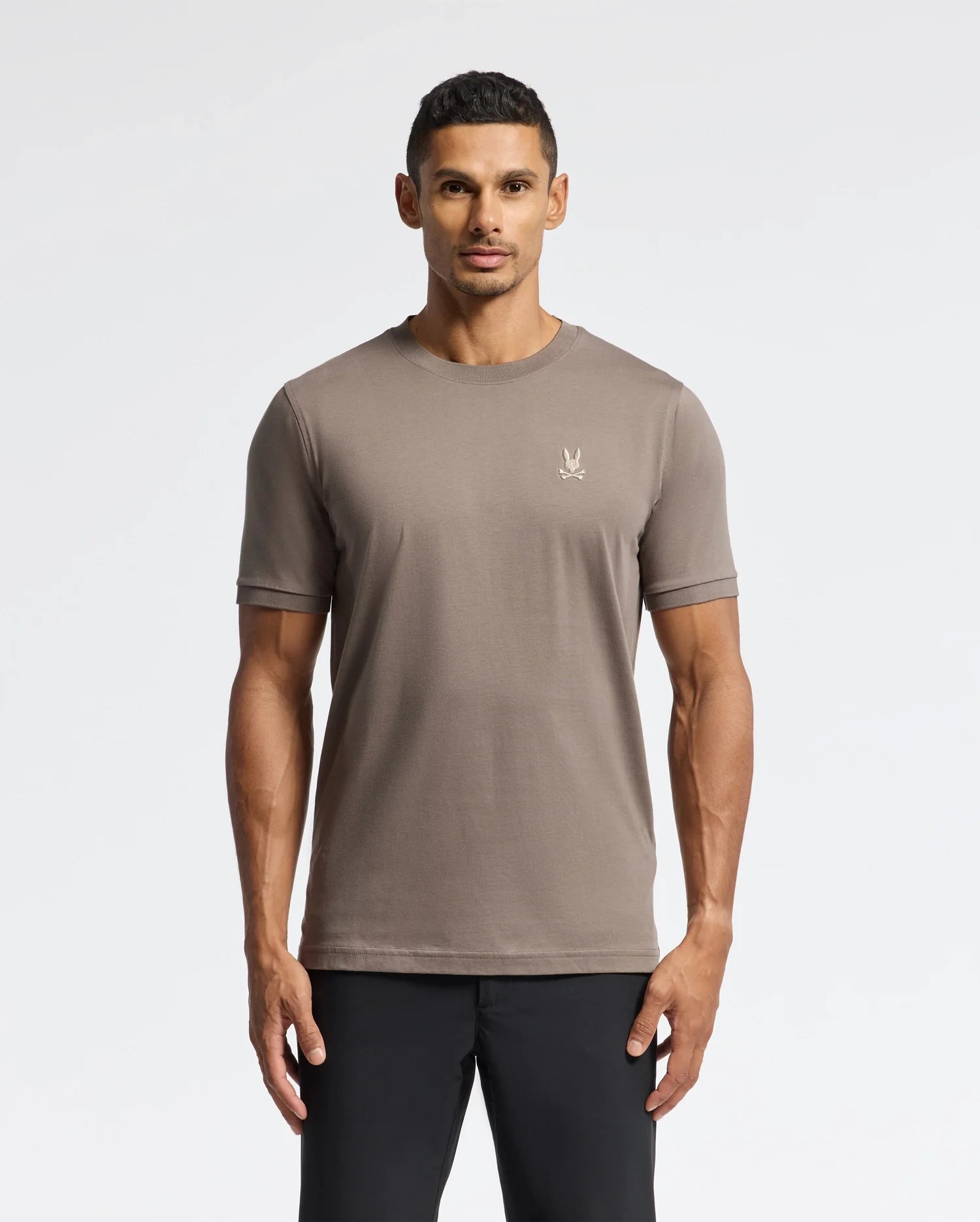 Against a plain white backdrop, a person wears the taupe Psycho Bunny MENS ALEXANDER FASHION TEE - B6U139E200 featuring a small embroidered logo on the chest. This luxurious tee embodies premium quality and timeless style, complementing their black pants perfectly.