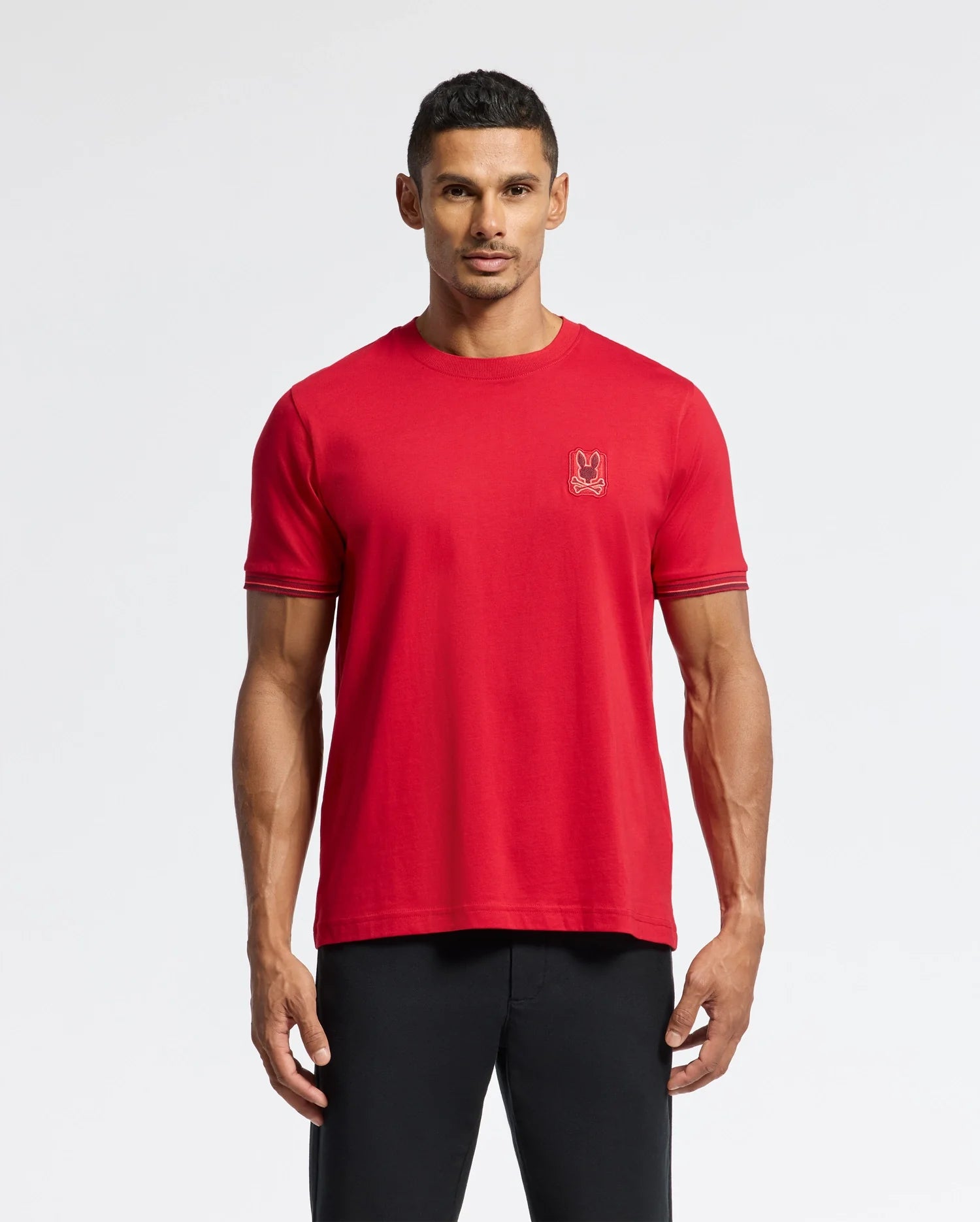 A man in a vibrant red Psycho Bunny Men's Pierce Fashion Tee, 100% Pima Cotton with dark sleeve trim and an embroidered logo on the chest, pairs it with black pants against a plain white background.