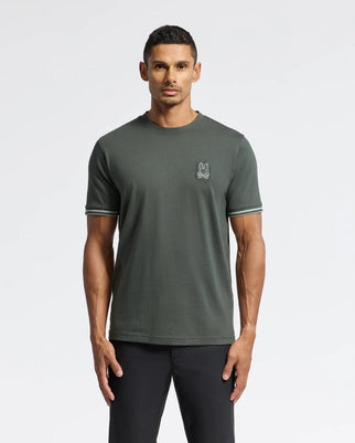 A person stands against a plain white background, wearing a dark green Mens Pierce Fashion Tee by Psycho Bunny, which features a small embroidered logo on the chest and dark pants. This premium T-shirt includes contrasting trim on the sleeves for a touch of casual elegance.