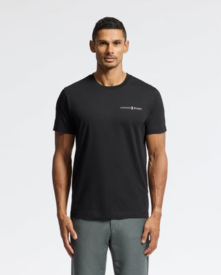 A man stands against a plain white background, clad in a premium Psycho Bunny top made from Pima cotton. The black MENS LONMONT GRAPHIC TEE - B6U194E200, which showcases a subtle chest logo, is artfully paired with gray pants. He sports short black hair and maintains a neutral expression.