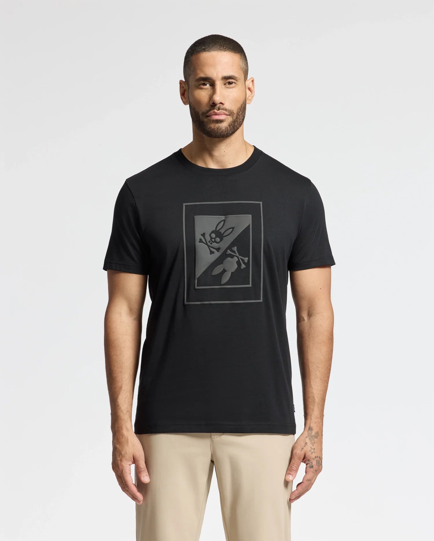 A man with short hair and a beard is wearing the trendy MENS DOTSER GRAPHIC TEE by Psycho Bunny, featuring a black butterfly design crossed by a diagonal line. He's paired it with beige pants to highlight his style, while his tattooed right hand adds an edgy touch against the plain white background.