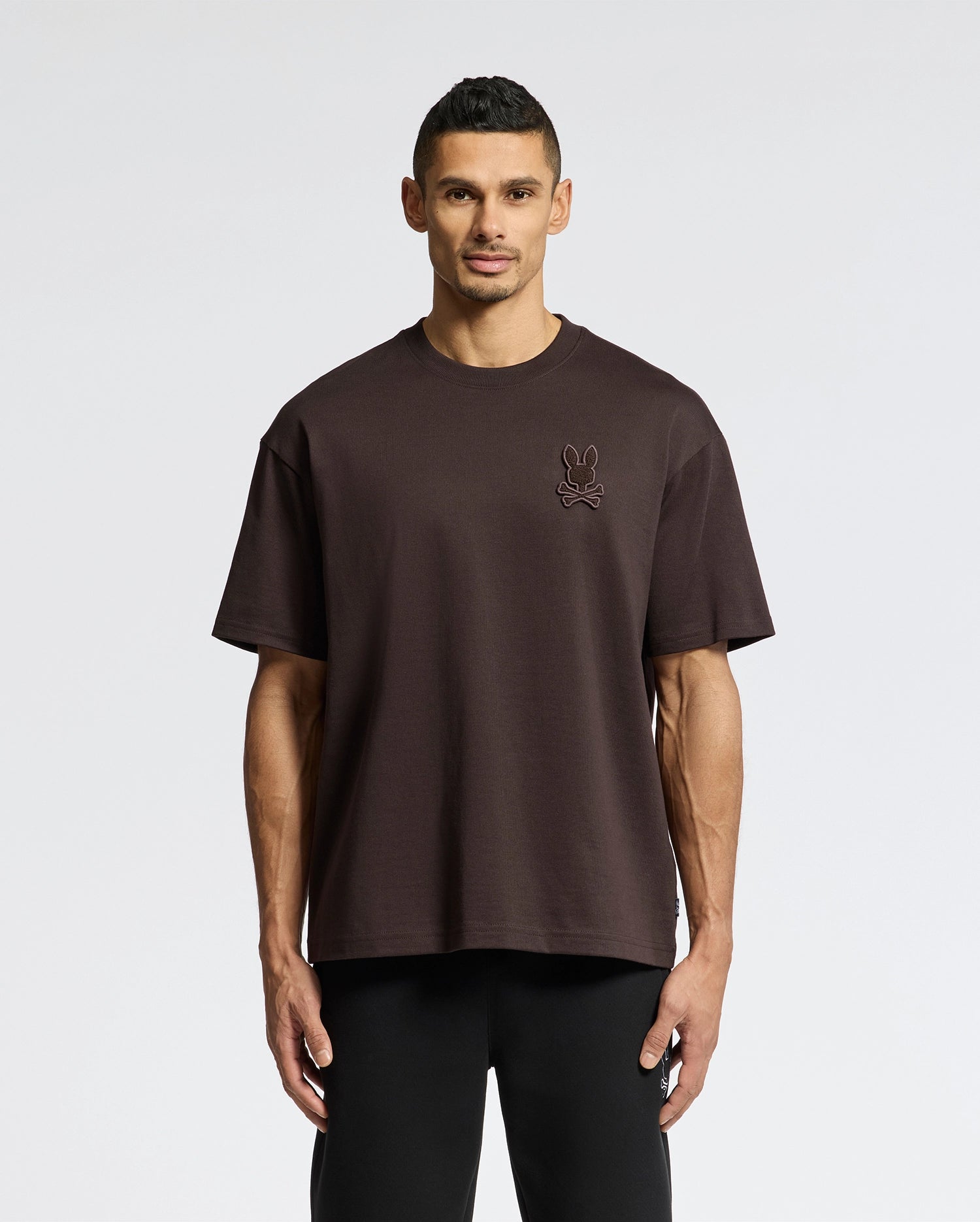 A man stands against a plain background, wearing a Psycho Bunny Men's Eldorado Heavy Weight Relaxed Fit Tee (B6U215E200) in dark brown Pima cotton with a small embroidered chest graphic, paired with black pants.