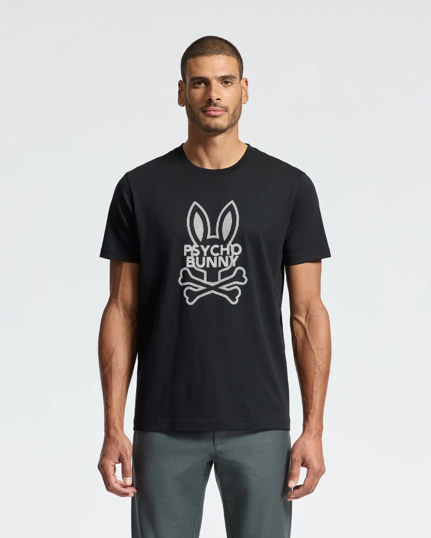 A man stands against a plain white background, sporting the MENS DUMON GRAPHIC TEE - B6U271E200 by Psycho Bunny. This premium t-shirt features a bunny skull and crossbones graphic with the text 