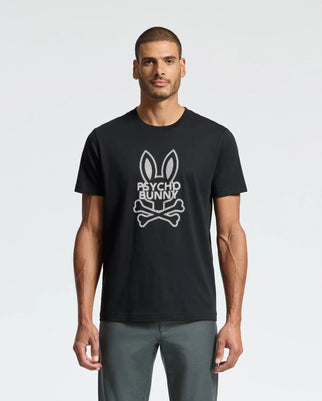 A man stands against a plain white background, sporting the MENS DUMON GRAPHIC TEE - B6U271E200 by Psycho Bunny. This premium t-shirt features a bunny skull and crossbones graphic with the text "Psycho Bunny" and is crafted from soft Pima cotton for both comfort and edgy flair.