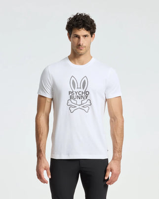 A man stands against a plain background wearing the MENS DUMON GRAPHIC TEE - B6U271E200 by Psycho Bunny, showcasing a stylized rabbit head with crossbones logo. Crafted from Premium Pima Cotton, he pairs the white graphic tee effortlessly with black pants.