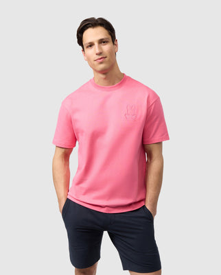 A man stands against a plain white background, wearing the bright pink MENS DANBY OVERSIZED TEE - B6U359B2TS and dark shorts. Made from relaxed-fit Heavyweight Pima cotton, the tee features an embroidered Bunny appliqué on the left chest by Psycho Bunny. His hands are casually resting in his shorts pockets as he smiles softly.