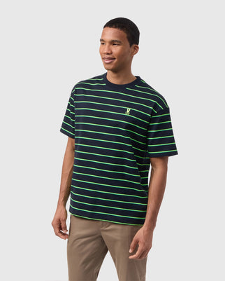 A man wearing a relaxed fit, black T-shirt with bright green horizontal stripes and light brown pants is smiling and looking to the side. The MENS ALTON STRIPE OVERSIZED TEE - B6U409C200 by Psycho Bunny, made of 100% Pima cotton heavyweight jersey, has a small embroidered logo on the left side of the chest. The background is plain and light-colored.