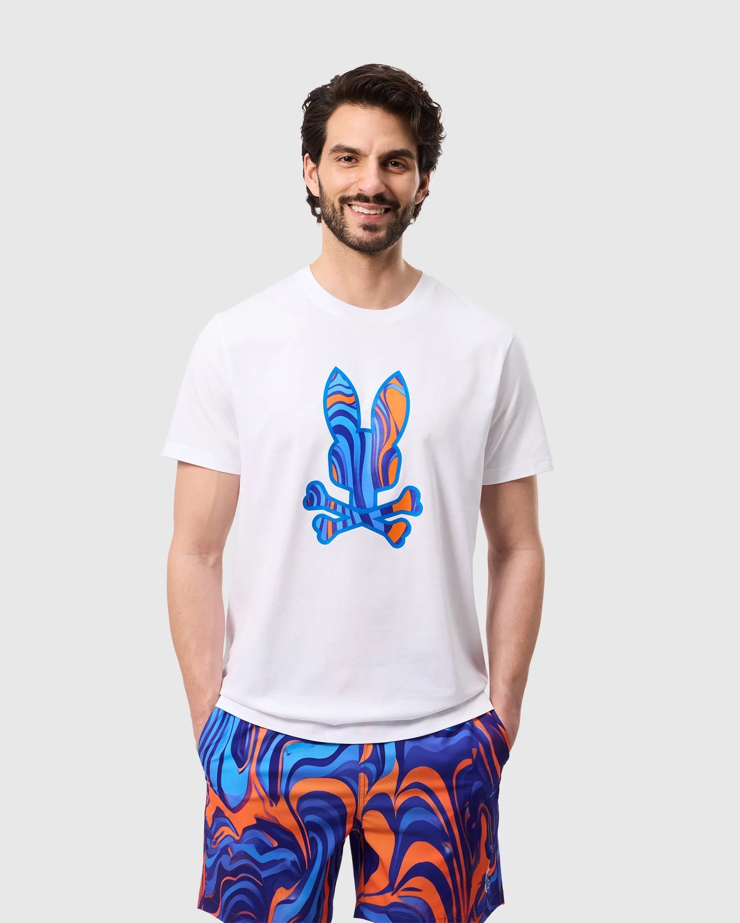 A man with dark hair and a beard is smiling, wearing a white Psycho Bunny MENS NEVADA GRAPHIC TEE - B6U515C200 featuring a colorful Bunny print and crossbones. He pairs it with brightly colored shorts showcasing a blue and orange swirl pattern. The background is plain light gray.

