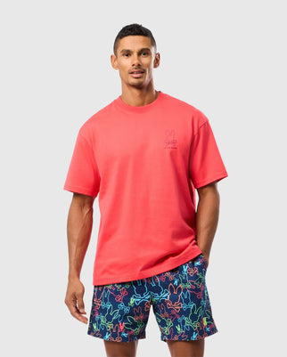 A man wearing the MENS BARRETT OVERSIZED TEE - B6U518C200 by Psycho Bunny, a relaxed fit bright red Pima cotton jersey t-shirt with a small bunny graphic on the chest, pairs it with colorful shorts featuring a playful abstract pattern while standing against a plain light gray background.