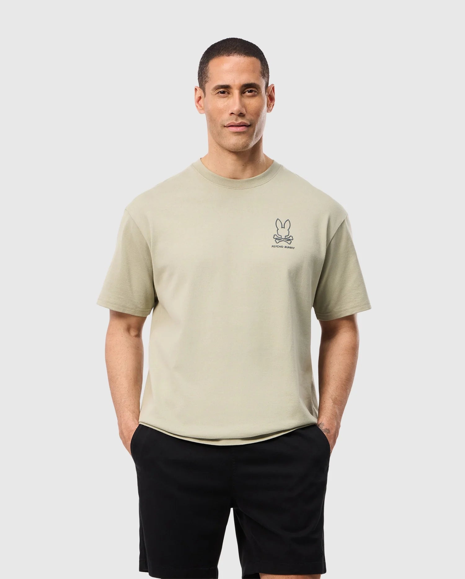 A person wearing the Psycho Bunny MENS BARRETT OVERSIZED TEE - B6U518C200, a beige Pima cotton jersey t-shirt featuring a small embroidered bunny graphic on the left chest, stands with hands in pockets. They also sport black shorts. The relaxed fit of the outfit contrasts nicely against the plain, light-colored background, making it stand out effortlessly.