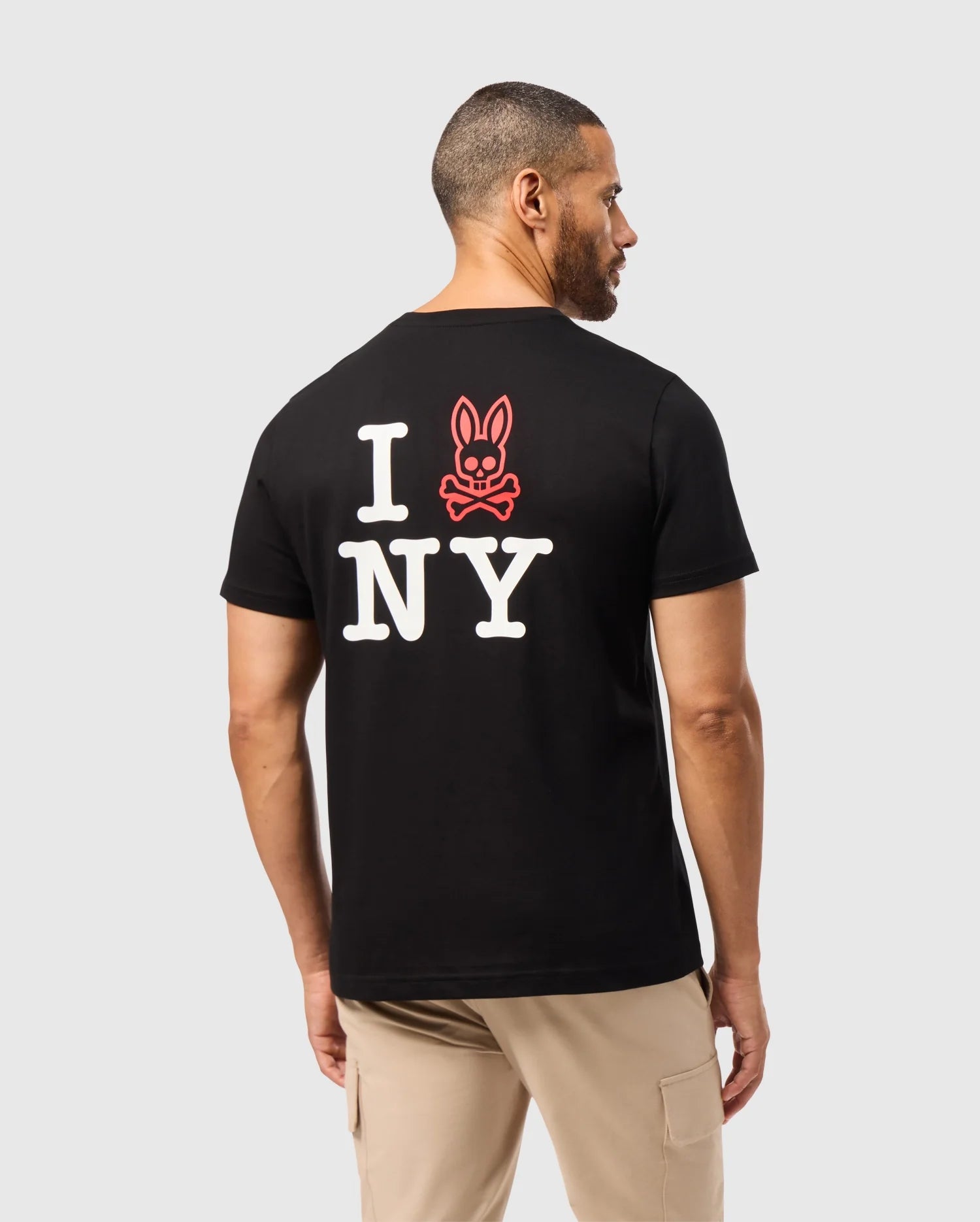 A man with short hair and a beard is wearing a Psycho Bunny MENS NEW YORK TEE - B6U554W1PC in black, featuring 