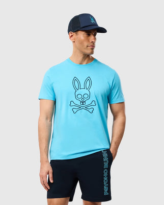 A man wears a Psycho Bunny MENS SHELDON GRAPHIC TEE in blue, depicting a bunny skull and crossbones on soft Pima cotton. He's paired it with black shorts that have side text and wears a black cap while standing against a plain gray background.